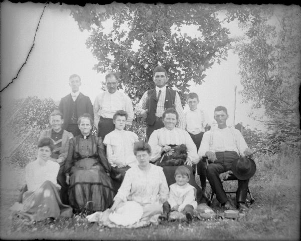 Nine unnamed members of the Haas family posing for an outdoor group portrait. They are joined by three Von der Sump sisters: Leta, sitting on the ground on the far left; Hattie on the ground in the center; and Mae (Maggie) sitting on the chair third from left.  