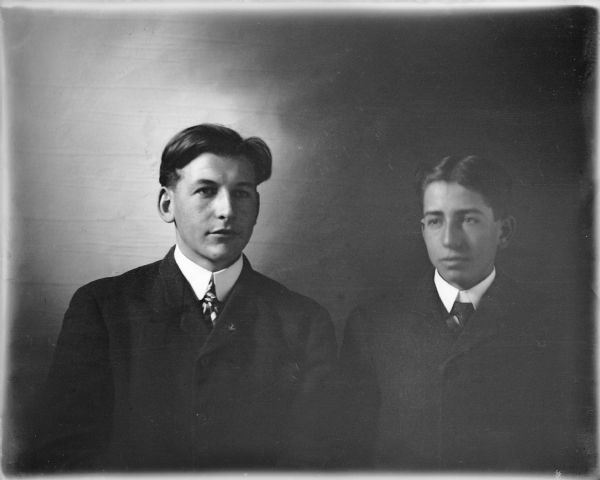 Fred von der Sump, left, posing with another young man for a head and shoulders portrait in front of a cloth backdrop. Both men are wearing suit coats and ties; Fred has a pin, possibly an anchor, on his lapel. A note on the negative envelope identifies the man on the right as Hobbart or Howard.