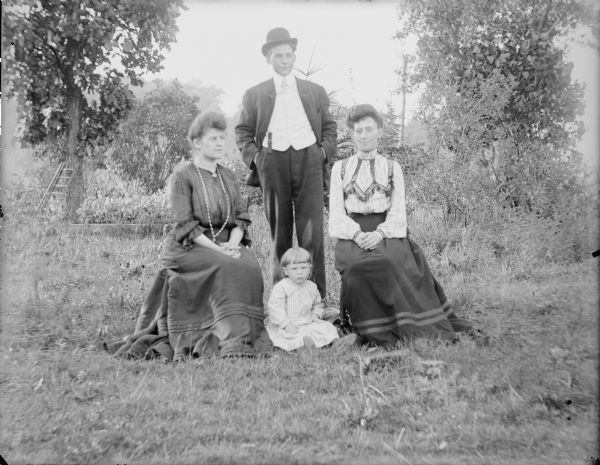 Hattie von der Sump, sitting at left, and her brother Fred, standing, posing with Mrs. Wallace and an unidentified toddler, who is sitting on the ground. The adults are very well-dressed. There is a ladder leaning against a tree in the background on the left.