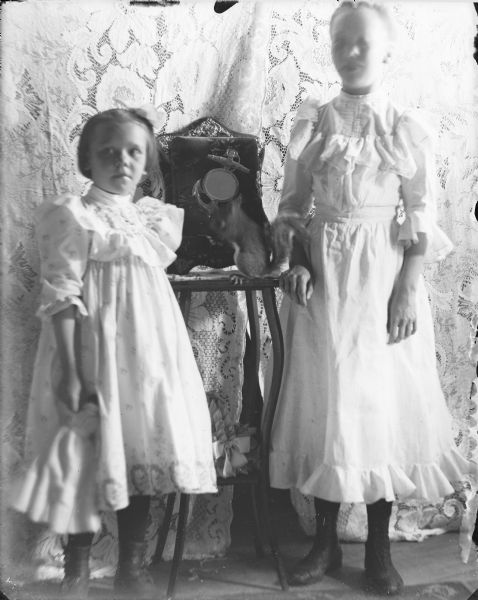 Two young girls wearing fancy dresses are standing on either side of a small table. The girl on the left is holding a doll in her right hand. There is a live squirrel sitting on the table in front of a photograph album propped against a lace cloth backdrop. The album has a velvet cover with an anchor and mirror decoration.