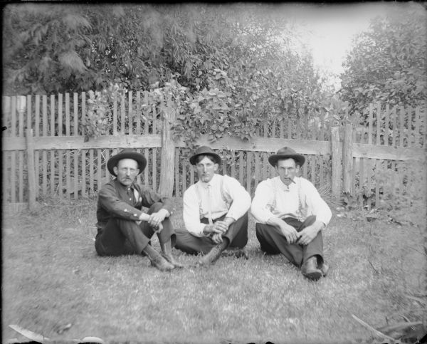 Three men are sitting on the ground in front of a picket fence. They are wearing hats and smoking cigars. The man at left appears to be older than the others. Written on the negative envelope is: "August, Frank and Fred Bernhagen."