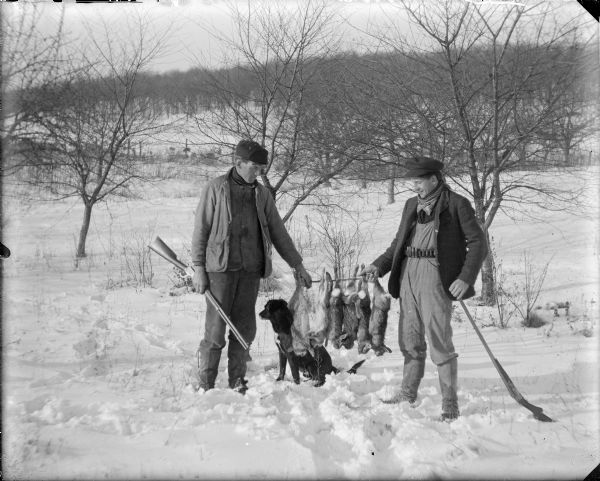 Will von der Sump, left, and his brother Fred are posing holding a brace of six rabbits on a stick between them. Each man is holding a shotgun and there is a dog sitting next to Will. The men are dressed similarly with hat, neck scarf, coat and gaiters. Fred is wearing a shotgun shell bandolier under his coat. There is snow on the ground.