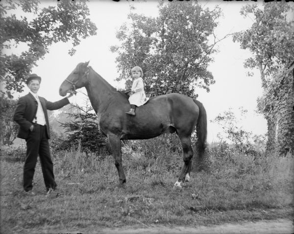 Fred von der Sump is holding the halter of a horse. A toddler identified as Jim and wearing a dress is sitting astride the horse bareback.