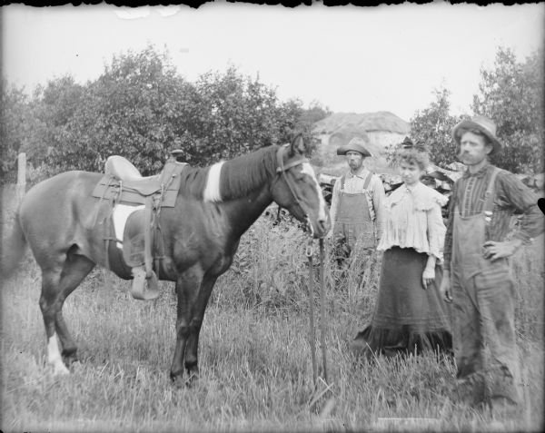 A horse with saddle and bridle is standing near a woman and two men. The woman is dressed in a fancy blouse and dress; the men are wearing bib overalls and hats. In the background is a hay or straw stack in what appears to be the foundation of a barn.