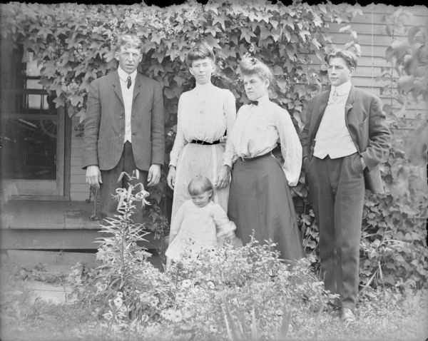 A group of four well-dressed adults, and a toddler wearing a dress, are posing in front of a vine-covered porch at the Von der Sump home. A lily and other flowers are blooming in front of the group. The four are, from left: Will von der Sump, Mrs. Wallace, Hattie and Fred von der Sump.  The child is not identified.