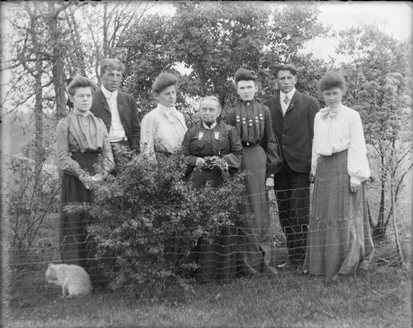 A group of five women and two men are posing behind a wire fence and shrub. They are, from left, Mae, Will and Hattie von der Sump; Mrs. Salisbury; a woman identified only as Edith; Fred and Leta von der Sump.  All are well-dressed. There is a cat at lower left.  