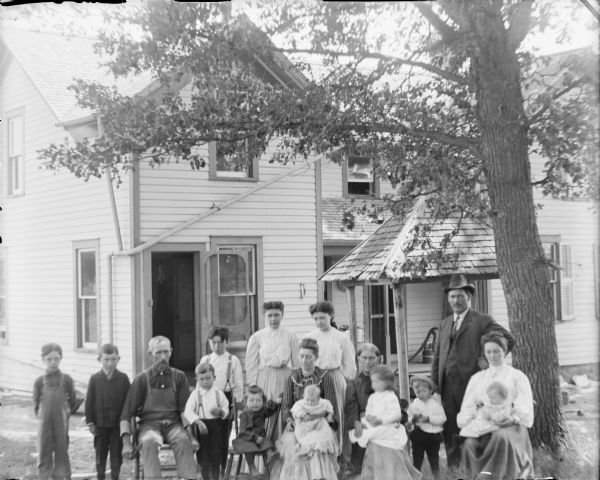 Group portrait of seven adults and nine children, including two infants, posing sitting and standing under a tree near the open door of a two-story wood frame house. There is an open, roofed structure at right over a hand pump.