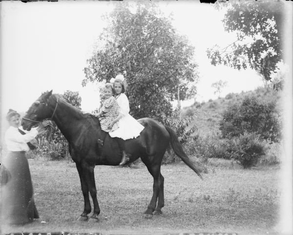 A woman at far left is holding the rope halter of a horse. A young girl and toddler are sitting bareback on the horse. The girl is wearing a light-colored dress and a bow in her hair. The toddler is wearing a plaid dress.