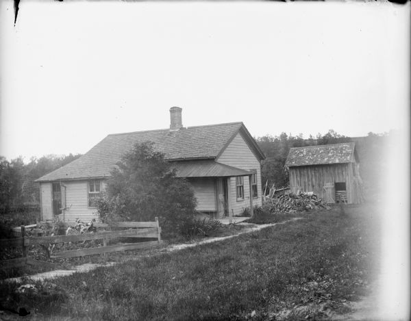 A board sidewalk leads to the porch of a modest one-story house. There is a lean-to style extension of the house to the left of the porch. In the background is a pile of scrap wood and a shed. A note on the negative envelope reads: "Our last house we lived in in Cambria."