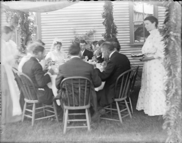 The bride, Hattie von der Sump, wearing her headpiece and veil is sitting at the far side of a round table next to groom Ellis Owens.  Other men and women are eating at the table, which  is set up on the lawn next to the wood sided Von der Sump house. Posts supporting a canopy above the table are decorated with fern fronds. Two women are standing at left, and another woman is on the right to serve the wedding party.