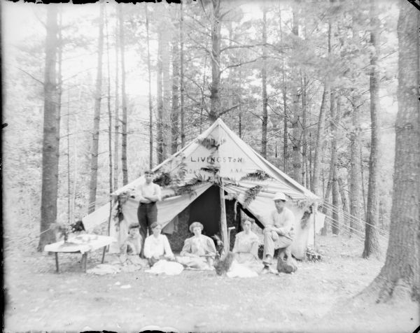 Four young women are posing sitting on the ground in front of a large tent. One man is standing behind the women at left, while another is sitting on a tree stump at right. Livingston, Montana has been printed on the tent above the door flaps, and fern fronds have been attached to the tent. There is a low table at left, with a tablecloth and a vase of flowers. Tall pines surround the tent.