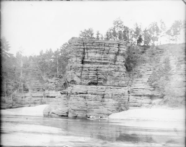 View across water towards people in a canoe on the Wisconsin River who are dwarfed by the High Rock formation at the Dells. There are sandy beaches on each side of the protruding point of High Rock. Trees are growing on the top of the cliff.