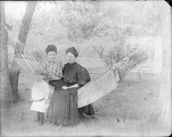 A little boy identified only as Clifford is standing next to two women who are sitting in a hammock. He is wearing a light-colored dress with a low belt. Leta von der Sump, in a plaid blouse, is sitting in the hammock on the left next to another young woman, Belle, who is wearing a dark dress.