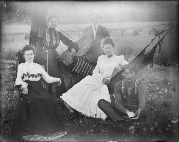 Five Von der Sump siblings posing for an outdoor portrait. Mae, at far left, is sitting in a chair with her feet on a pillow. Behind her is Leta, with an arm resting on the bar of a hammock. Will is at center rear. Hattie is sitting in the hammock holding a newspaper, and Fred is sitting on the ground, also holding a paper or pamphlet. There are wheat shocks in the field behind them.