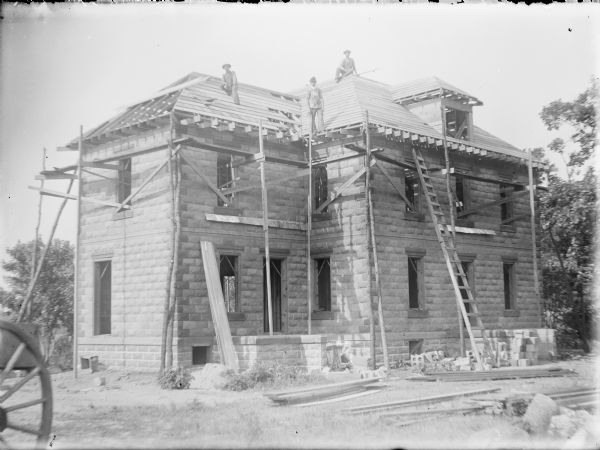Three men, identified on the negative envelope as "Mr. Miller, Boots, and Lavine," are posing on the roof of a large two-story cement block house under construction. The doors and windows have not been installed. There is scaffolding built of planks and narrow tree trunks anchored to the window openings.