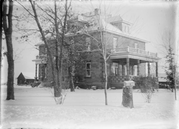 A winter view of the front and one side of the newly completed cement block house of Frank and Bertha von der Sump on Golden Road. The four square style house has an extension on the rear and two dormers are visible. Surrounding the central chimney is a widow's walk with low fence. There is a large front porch and two side porches. A mailbox is on a stump at the edge of the road. There is snow on the ground and farm buildings in the background.