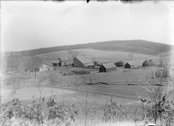 Elevated view from hill of the Frank and Bertha von der Sump farm on Golden Road. There is a modest one-story farmhouse, a barn, windmill, and four other farm buildings with haystacks below. There is a summer kitchen behind the house and an outhouse to the right, near two woodpiles. Cows are penned beside the barn and chickens range free. A small carriage is parked near the house. Fields surround the farmyard, and wooded hillsides are in the distance.