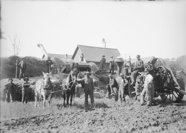 Thirteen men take time from threshing duties to pose for a group portrait with a team of horses, threshing machine with straw elevator, and a steam tractor amid stacks of wheat. Some of the men are holding pitchforks. Farm buildings and a windmill are in the background.