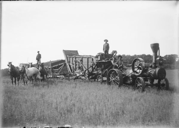 A man at left is standing and holding the reins of a four horse hitch, while three other men are posing between a steam tractor and threshing machine. There is a straw elevator at the rear. Wheels and belts are easily seen on the side of the thresher.
