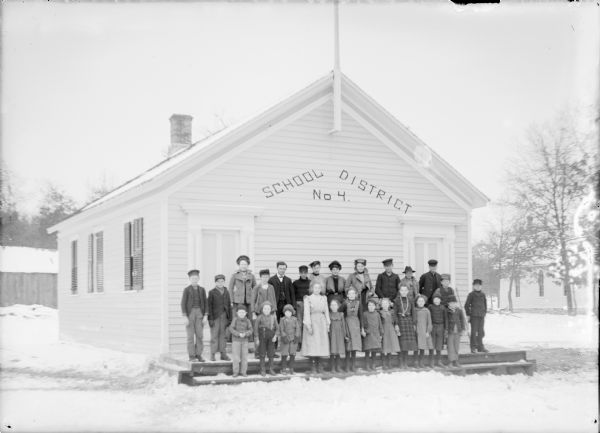 Group portrait of children and adults standing on the open front porch of a small wood frame school building with two front doors. Among the adults in the second row, fifth from left, is a well-dressed man wearing a suit and bow tie who may be the teacher. Painted on the front of the school is "School District No. 4." There is a small church building in the background on the right. A notation on the negative envelope identifies this as "Valley School House."