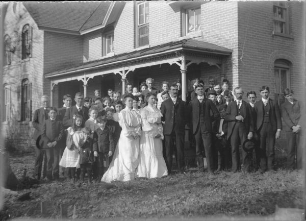 Group portrait, with bride and groom Anna Kearns and Neil J. Brown, (with moustache) standing at the center front on their wedding day. They are flanked by their attendants, with family and guests gathered behind them in front of a two-story brick house.