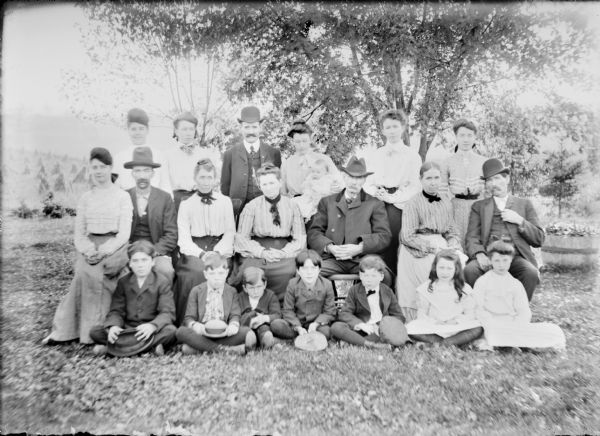 Seven children are sitting on the ground in front of a row of seven adults who are sitting, with six adults standing in the rear. A woman in the back row, center, is holding an infant. Notes on the negative envelope state: "Group of Tom Kearns, T. [Thomas] Mahar, Pl [Peter] McMahon and Tom Duffy and family." The group is well-dressed. There are wheat or rye sheaves in a hillside field at left.