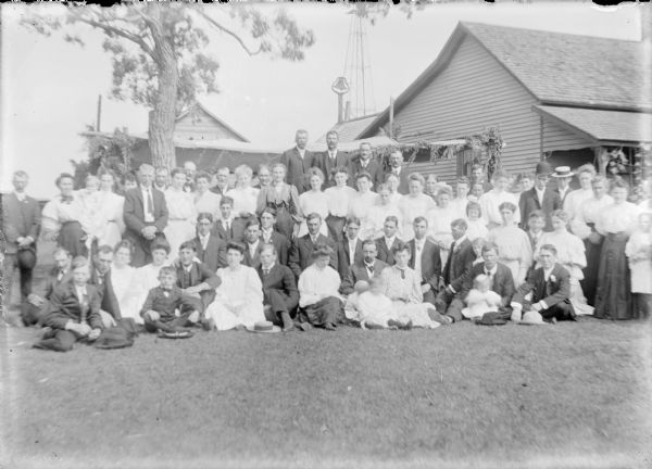 A large group of well-dressed men, women and children posing beside the small wood frame Von der Sump farmhouse. There is a canopy decorated with greenery behind the group. A dinner bell and windmill tower are in the background.  The bride, Hattie von der Sump, and groom, Ellis Owens, are seen standing at far right.  He is wearing a derby hat and Hattie is to the left.