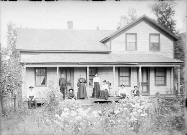 A well-dressed man and woman are standing between two columns on the front porch of a wood frame house. There are two younger women at right, with one sitting in a chair. Three men and a boy (far right) are sitting on the porch floor. There is a dog next to the boy. Flowering plants are in the front yard in the foreground.