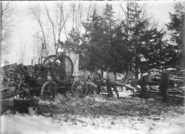 Will von der Sump, second from left, is watching as two men guide a log into the circular saw blade of a portable sawmill. A steam engine at left powers the saw. Two other men are posing at right at the base of a pile of logs. Fire wood is stacked at left. Snow is on the ground.