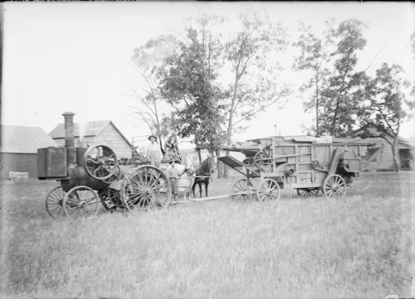 A man in bib overalls is standing on the rear of a steam tractor which is hitched to a mechanical huller. Behind the tractor is a wagon full of firewood drawn by a team of horses. One man is holding the bridle of one of the horses while another man is sitting on a barrel in the wagon. In the background is a small farmhouse on the right, a windmill behind a tree, and other farm buildings on the left. A note on the negative envelope states: "A. Davis and huller outfit."