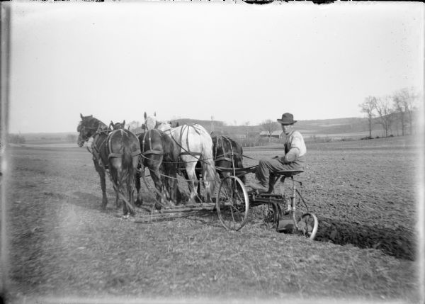 Will von der Sump driving a team of four horses from the seat of what appears to be a single bottom sulky plow. He is in a large field on his family's farm; the farm buildings and windmill are in the background. A note on the negative envelope identifies the implement as a gang plow.