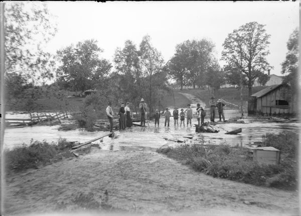 Water from French Creek covers the surface of a bridge at Dates Mill near the Marquette-Columbia County line as water rushes down a lane in the pasture in the background. Several men, boys, and two women are on the bridge. The caption on the negative envelope states: "Mae, Leta [Von der Sump] and the boys on the bridge at Dates." There is a shed at right and cows in the background at left. It appears that a portion of the bridge has been swept away, and there is a damaged fence at left.