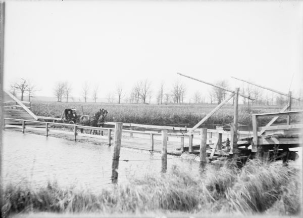 View from shoreline looking towards two women wearing large hats who are sitting in a carriage drawn by a matched team of horses. The carriage has stopped on a float bridge across the Fox River. The plank road surface of the bridge is barely above water level. The riverbanks are covered with marsh grasses, and there are trees in the distance.