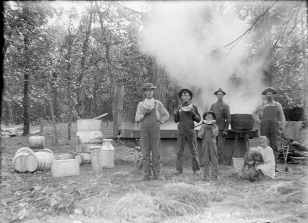 Steam is rising from a "cooking pan" where sorghum juice is boiling. There is a smokestack at left and a pile of fire wood on the right. Barrels, milk cans, and wash tubs are at left. Two young men and a boy are eating watermelon in front of the pan; two men are standing behind them. A young girl at right is sitting on a bucket while holding a dog.