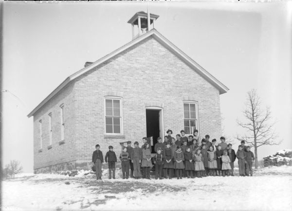 Children of a range of ages are standing with their teacher in front of the Browning School. The children are well-dressed in winter clothes. The school is built of light-colored brick and has a dressed stone foundation. There is a bell tower in front and a chimney at the rear. A wood pile is on the far right.