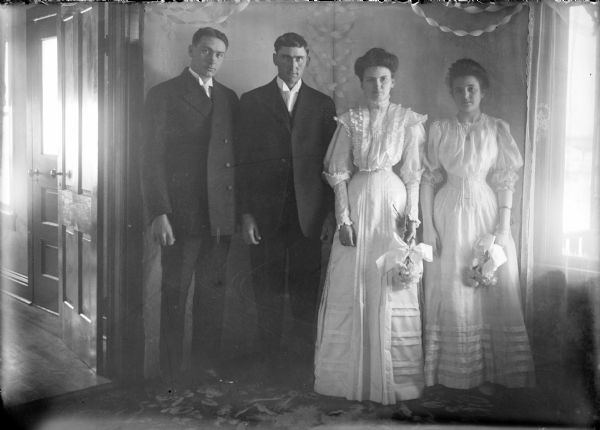 An interior, full-length standing portrait of a bride, Edith Thompson, and groom Herman Kamrath, flanked by their attendants. The women are wearing similar long dresses with narrow waists, and are holding bouquets in their left hands. The men are wearing dark suits and white ties. There are crepe paper streamers overhead and in the corner behind them.