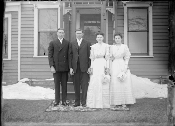Outdoor wedding portrait of Edith Thompson, second from right, and groom Herman Kamrath. They are flanked by their attendants. The group is standing on a small carpet on the ground in front of a wood sided house. The women are wearing similar light-colored dresses with cinched waists, and they are both holding a bouquet in their left hands. The men are wearing dark suits and white ties. There are piles of snow near the foundation of the house.