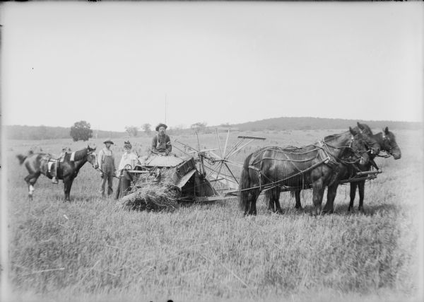 A woman holding the reins of a saddled horse is standing next to a Milwaukee No. 10 grain harvester and binder hitched to three horses. A man is sitting on the seat of the binder, and another man is standing behind the woman on the left. The hitched horses have fly-nets. Wheat shocks are in the field in the background.