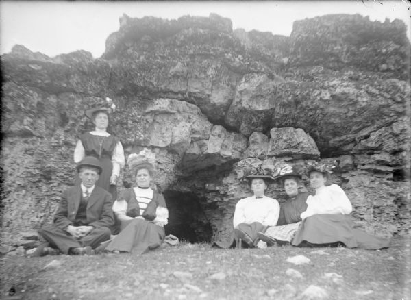 Will von der Sump, far left, is sitting on the ground in front of his sister Mae and next to his sister Hattie in front of a large rock outcropping. Their sister Leta is sitting between two unidentified women at right. Between the two groups is what appears to be the opening of a cave. The group is well-dressed and all of the women are wearing large hats.