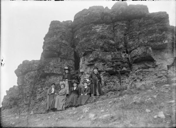 A group of five women and a man are posing in front of a large rock outcropping. The man who is standing at right is unidentified. Leta von der Sump is standing behind four women who are sitting on a rock ledge. They include Hattie von der Sump, second from left, and Mae von der Sump, fourth from left. The other two women are unidentified. The women are wearing overcoats and large hats.