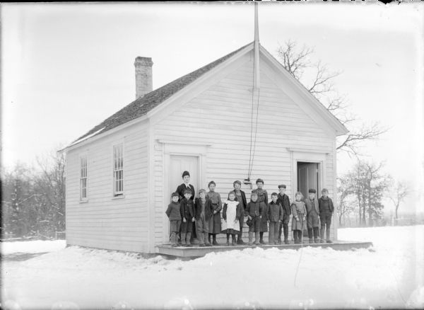 Leta von der Sump, standing at rear left, posing with thirteen children on the front platform of Belle Fountain School. There are two doors on the front of the wood frame building. A flagpole is mounted in the front gable, and there is a brick chimney at the rear. Snow is on the ground.