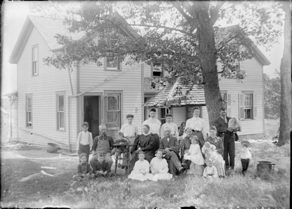 Group portrait of three men, seven women, and eleven children, including two infants, posing in front of a two-story wood frame house. They are shaded by two large trees. A small child is sitting in a high chair. An open structure with a shingled roof over a water pump is just behind the group.