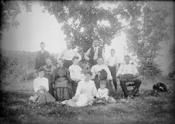 An outdoor group portrait of members of the Haas and Von der Sump families. They include Leta von der Sump, sitting on the ground on the   far left; Hattie von der Sump, on the ground in center, and their sister Mae, sitting on a chair behind and to the left of Hattie. The others are not identified. There is a dog in the grass on the far right.