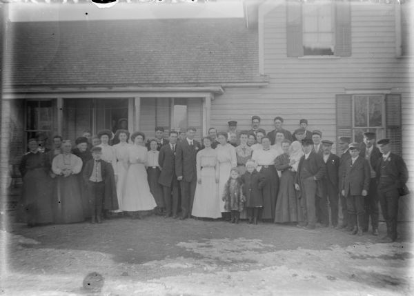 A large group of well-dressed men, women and children are posing in front of a wood frame house. At the center of the group is a man with a boutonniere, presumably the groom, standing next to his bride who is wearing a light-colored dress. They are flanked by a similarly dressed couple. There is snow on the ground and many of the men and boys are wearing hats. Two children standing in front of the wedding party are wearing heavy coats.