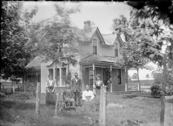 A man and woman are posing sitting on chairs on the lawn of a wood frame farmhouse. A man and two young women stand behind them. The group is well-dressed. They are identified on the negative envelope only as "Lampke Family."