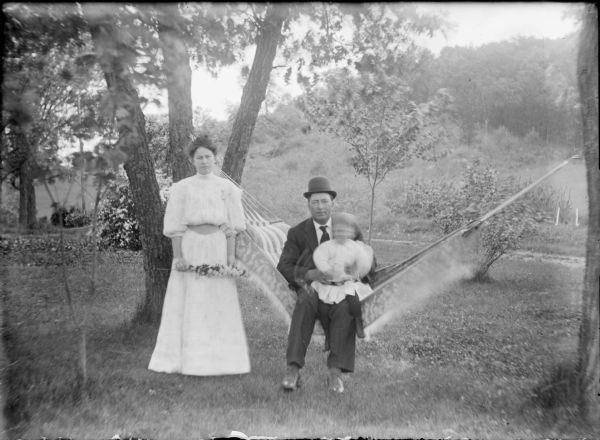 A man wearing a suit and hat is sitting on a hammock holding a small child (blurred by movement) in his lap. Standing at left is a woman wearing a fancy light-colored dress who is holding a stem of flowers. A locket or watch is pinned to her dress. They are on the lawn of the Von der Sump farmhouse. A note on the negative envelope identifies the family as "Dave O'Keefe, Belle and Baby."