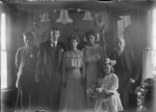 An available light interior group portrait of a bride and groom with their attendants, an older gentleman, likely the officiant, at far right and a flower girl in front. The bride's attendant and the flower girl have large bows in their hair. The men all wear suits and ties. Paper honeycomb bells and other decorations hang from the ceiling; there is a parlor stove in the right foreground.