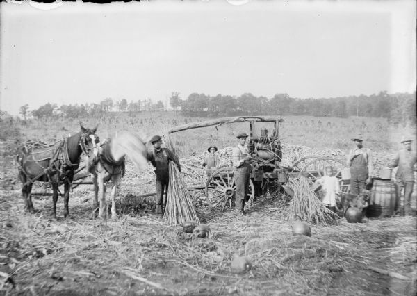 A little girl posing with men and boys, probably members of the Cuff family, around a portable sorghum mill. One young man is holding a bundle of cane as another man feeds cane into the crusher. The mill is powered by two horses (at left) hitched to a long log mounted to the top of the mill. At right, two men and a dog are standing near a wooden barrel. A young boy in the background is holding a long handled tool, probably a pitch fork to clear the crushed cane from the mill. There are pumpkins scattered on the ground, and a field of corn shocks in the background.