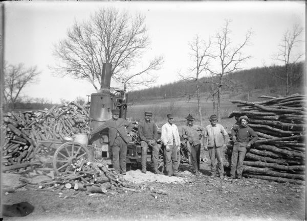 Six men posing in front of a portable steam driven circular saw on the Von der Sump farm. A portion of the saw blade is visible between two men in the center. There is a pile of stove wood behind the saw and a stack of logs at right. In the background is a wooded hillside.