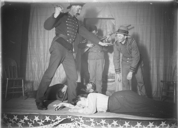 In a scene from a play, a woman is lying on the stage floor with her eyes closed. Two men in military uniforms, one brandishing a sword, are standing over her. Another actor behind them attends to his gun as a man in civilian dress reclines on the floor. There is patriotic bunting hanging at the front of the stage. A note on the negative envelope states: "Edith V. fainted. Henry P. Wallace [illegible] dares to touch her."